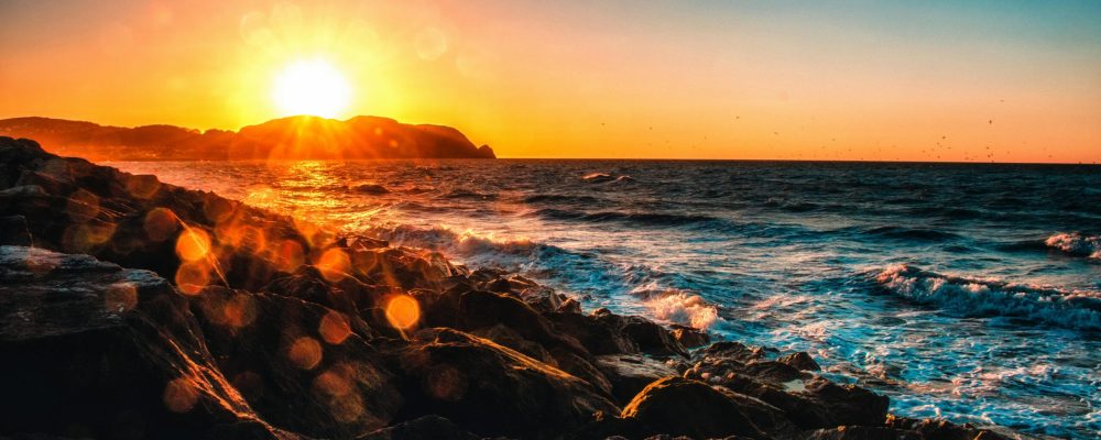Captivating sunset view over a rocky seashore with vibrant sun glare and waves crashing.